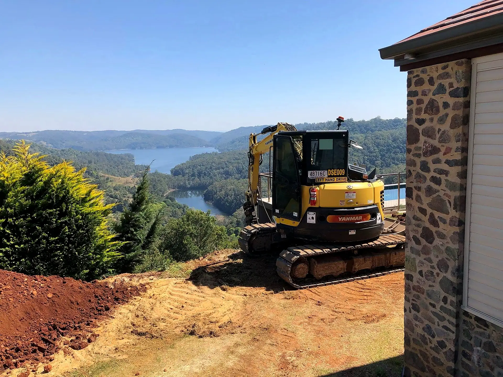 Milako's yellow excavator on a freshly cut hillside pad beside a stone house, with a lake and valley behind